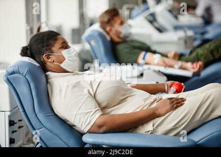 Seitenansicht Porträt von Menschen, die Masken tragen, während sie im medizinischen Spendenzentrum Blut in Reihe geben Stockfoto