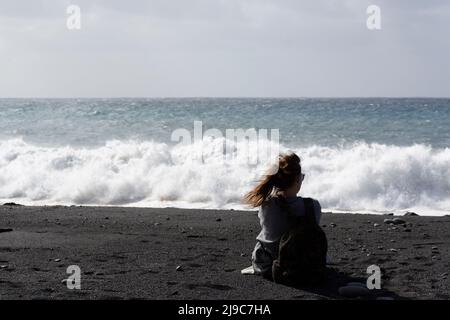 Eine nicht identifizierte junge Erwachsene Frau, die auf ozeanischem stürmischem Wasser und Steinen am schwarzen Lavastrand in Tazacorte auf der Insel La Palma, Kanarische Inseln, Sp Stockfoto