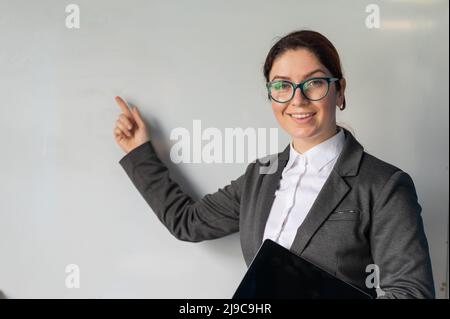 Schöne rothaarige Frau in einem Anzug mit einem digitalen Tablet in den Händen. Eine lächelnde Lehrerin mit Brille steht in einer Schulklasse und zeigt ein Stockfoto