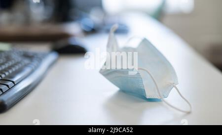 Büromitarbeiter-Desktop während einer Pandemie. Aufnahme einer verwendeten Maske durch die Tastatur auf einem weißen Tisch. Der Manager nahm die gebrauchte Maske am Arbeitsplatz ab Stockfoto