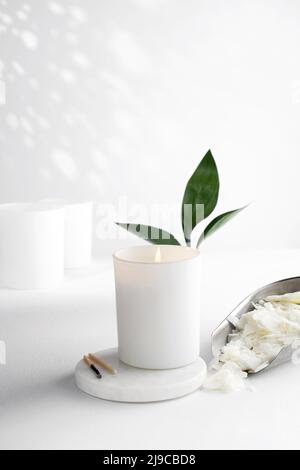 Sojawachskerze in einem weißen Keramikglas und grünes Blatt, Pflanze auf weißem Hintergrund. Natürliche umweltfreundliche Bio-Wachskerzen. Trendiges Konzept. Stockfoto