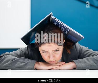 Probleme bei der Arbeit. Eine erschöpfte Frau faltete ihren Laptop über ihren Kopf an ihrem Schreibtisch. Geschäftsfrau in Frustration unter ihrem Computer begraben. Reduzierung Stockfoto