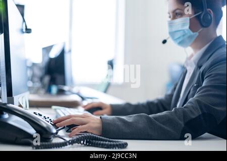 Eine Frau, die eine medizinische Maske in einem Freiraumbüro trägt. Eine Sekretärin nimmt Anrufe von Headset-Kunden entgegen. Soziale Distanz am Arbeitsplatz. Hygienestandards Stockfoto