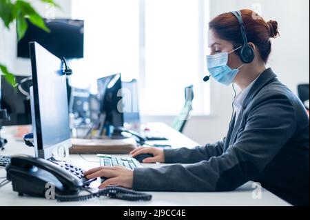 Eine Frau, die eine medizinische Maske in einem Freiraumbüro trägt. Eine Sekretärin nimmt Anrufe von Headset-Kunden entgegen. Soziale Distanz am Arbeitsplatz. Hygienestandards Stockfoto