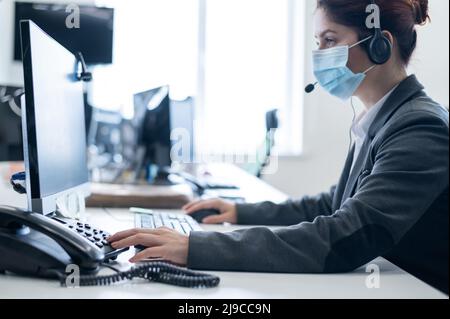 Eine Frau, die eine medizinische Maske in einem Freiraumbüro trägt. Eine Sekretärin nimmt Anrufe von Headset-Kunden entgegen. Soziale Distanz am Arbeitsplatz. Hygienestandards Stockfoto