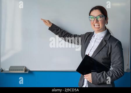 Verärgerte Geschäftsfrau Trainer zeigt Finger auf Whiteboard im Büro.Eine verärgerte weibliche Chef hält eine Tablette und schreit auf Untergebene in der Stockfoto
