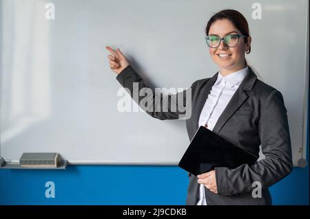 Schöne rothaarige Frau in einem Anzug mit einem digitalen Tablet in den Händen. Eine lächelnde Lehrerin mit Brille steht in einer Schulklasse und zeigt ein Stockfoto