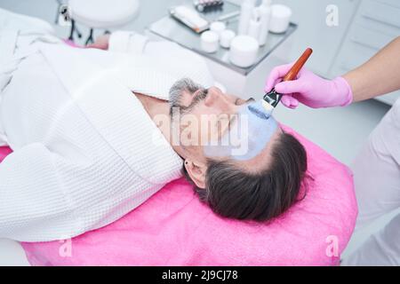 Weibliche Kosmetologin, die eine blaue Gesichtsmaske auf das Gesicht eines erwachsenen Mannes mit Bart aufträgt Stockfoto