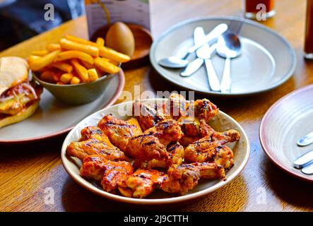 Gegrillte Hähnchenflügel auf dem Esstisch des Restaurants, mit unscharfen Chips, Messern und Tellern im Hintergrund Stockfoto