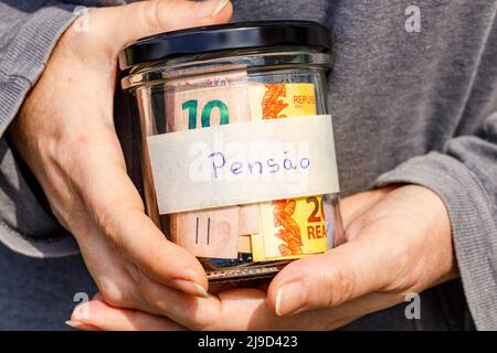 Eine Frau, die ein Glas mit Rentengeld in den Händen hält, eine handschriftliche Inschrift auf dem Glas Ruhestand in Portugiesisch, brasilianisches Geld, Konzept von p Stockfoto