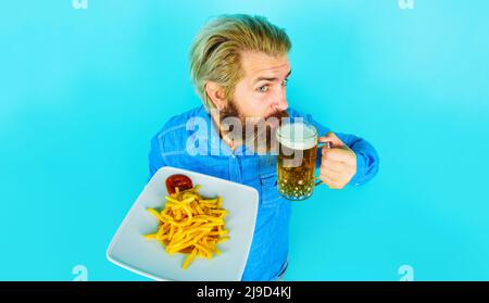 Bärtiger Mann, der Pommes isst und Bier trinkt. Gebratene Kartoffeln und Ketchup auf dem Teller. Fast Food. Stockfoto