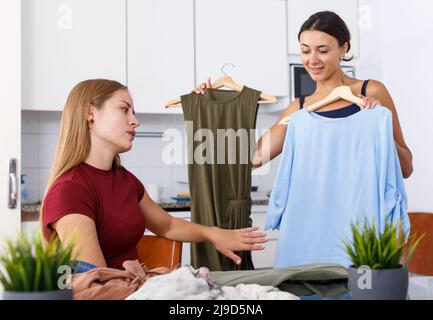Zwei junge positive Freundinnen suchen neue Kleider am Tisch und Freigabe in Home Stockfoto