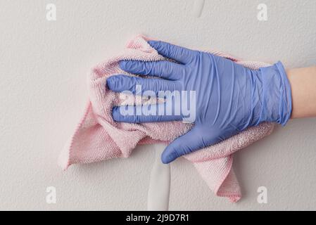 Die Hand des Mitarbeiters in einem blauen Gummischutzhandschuh mit Mikrofasertuch, das die Wand vor Staub abwischt. Zimmermädchen oder Hausfrau kümmert sich um Haus. Gewerbliche Reinigung Stockfoto