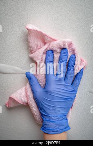 Die Hand des Mitarbeiters in einem blauen Gummischutzhandschuh mit Mikrofasertuch, das die Wand vor Staub abwischt. Zimmermädchen oder Hausfrau kümmert sich um Haus. Gewerbliche Reinigung Stockfoto