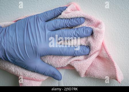 Die Hand des Mitarbeiters in einem blauen Gummischutzhandschuh mit Mikrofasertuch, das die Wand vor Staub abwischt. Zimmermädchen oder Hausfrau kümmert sich um Haus. Gewerbliche Reinigung Stockfoto