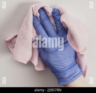 Die Hand des Mitarbeiters in einem blauen Gummischutzhandschuh mit Mikrofasertuch, das die Wand vor Staub abwischt. Zimmermädchen oder Hausfrau kümmert sich um Haus. Gewerbliche Reinigung Stockfoto