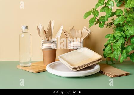 Set aus Einweggeschirr und Utensilien. Gabeln und Messer aus Holz, Pappbecher und Teller auf beigefarbenem grünen Hintergrund. Umweltfreundliches Geschirr. Stockfoto