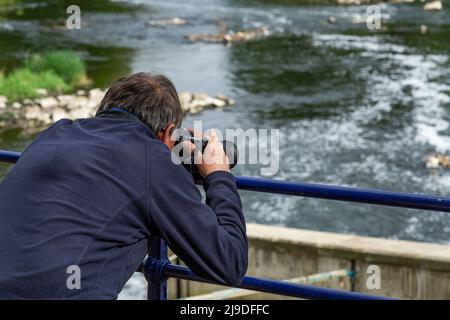 Ein männlicher Fotograf lehnt sich über Geländer, um ein Bild vom Fluss Aire in Saltaire, Yorkshire, zu machen. Stockfoto