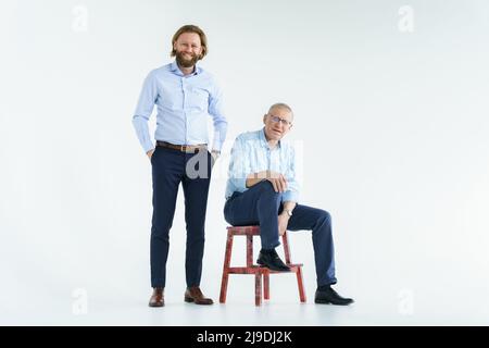 Vater sitzt auf dem Stuhl und Sohn steht auf weißem Hintergrund, beide Männer blicken in die Kamera, ein älterer Mann in Brille mit Dioptrien, ein stilvoller Junge Stockfoto