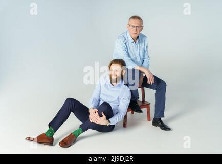 Vater sitzt auf dem Stuhl und Sohn sitzt auf weißem Hintergrund, beide Männer blicken in die Kamera, ein älterer Mann in einer Brille mit Dioptrien, ein stilvoller junger Mann Stockfoto