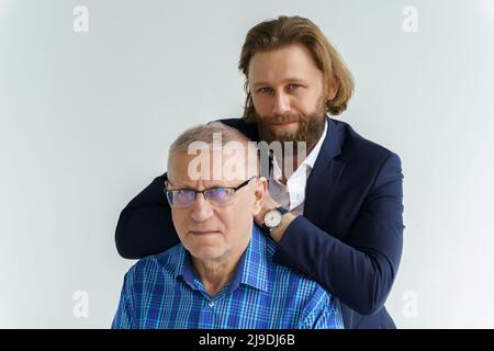 Der Sohn im stylischen Anzug lehnt sich auf die Schultern seines Vaters, Vater und Sohn auf weißem Hintergrund, beide Männer blicken in die Kamera, ein älterer Mann ist dabei Stockfoto