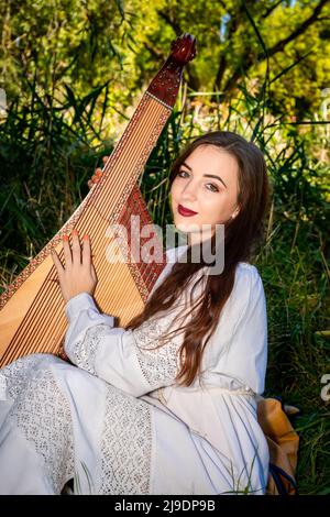 Die junge Ukrainerin in einem weißen Kleid und mit fließendem Haar sitzt auf dem grünen Gras und hält eine Bandura. Stockfoto