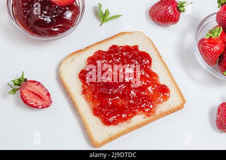 Toast mit Erdbeermarmelade zum Frühstück auf weißem Hintergrund. Draufsicht. Frische Erdbeeren und hausgemachte Erdbeermarmelade reifen, auf Brot verteilen. Stockfoto