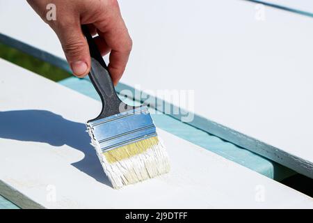 Männlicher Baumeister malt weiße Bretter für den Bau. Die männliche Hand hält die Bürste. Lackarbeiten, Hausrenovierung. Stockfoto