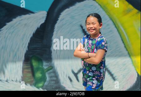 Portrait von asiatischen niedlichen Kind Mädchen auf der Straße mit kostenlosen Cartoon Graffiti Hintergrund für Touristen, ein Bild zu machen, kleines Mädchen mit großen Lächeln, Arme cr Stockfoto