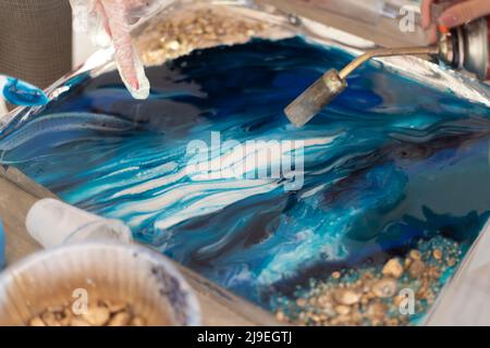 Trocknungsfarbe in blauer Farbe mit einem Haartrockner auf dem Brett. Weibliche Hand hält einen manuellen Gasbrenner, erstellen Sie ein Gemälde in Kunstharztechnik Stockfoto