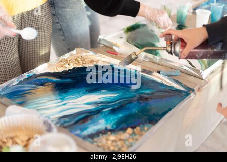 Trocknungsfarbe in blauer Farbe mit einem Haartrockner auf dem Brett. Weibliche Hand hält einen manuellen Gasbrenner, erstellen Sie ein Gemälde in Kunstharztechnik Stockfoto