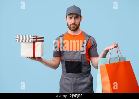 Trauriger männlicher Kurier in Uniform, der Einkaufstaschen und Geschenkkartons auf blauem Studiohintergrund hält Stockfoto