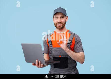 Gutaussehender lächelnder Mann in Uniform mit Laptop, der die Kamera ansah und ein Okey-Zeichen zeigte Stockfoto