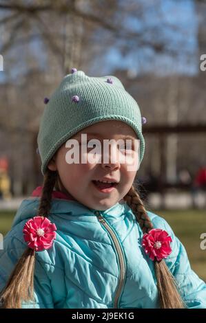 Porträt eines Mädchens mit langen braunen Haaren, die in Zöpfe geflochten sind. Ein Kind in einer Jacke und einem Hut blickt an der Kamera vorbei. Tagsüber. Frühling. Stockfoto