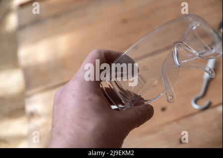 Eine Erwachsene männliche Hand hält die Schutzbrille. Metallschraube mit Dübel ruht auf einer hölzernen Oberfläche. Schraube mit C-Haken. Sicherheit am Arbeitsplatz, PSA-Konzept. Wählen Sie Stockfoto