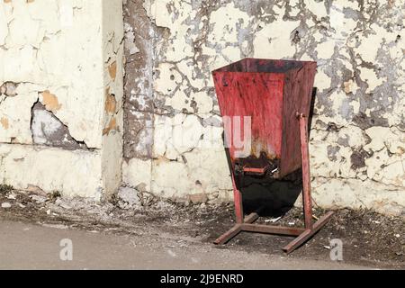 Der rostige rote Abfalleimer steht neben der schmucklosen alten Betonwand Stockfoto