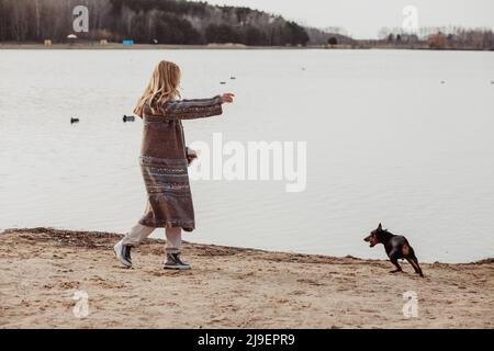 Glücklich genießen blonde Frau in warmen Kleidern spielen und Spaß mit niedlichen Welpen in der Nähe Flussufer an kühlen sonnigen Tag, Enten schwimmen im Wasser Stockfoto