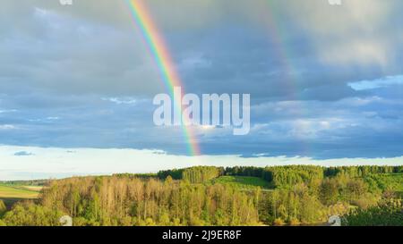 Doppelter Regenbogen als seltenes Naturphänomen vor dem Hintergrund einer hügeligen ländlichen Landschaft. Heller realer Regenbogen über dem Wald im frühen Frühjahr. Der Stockfoto