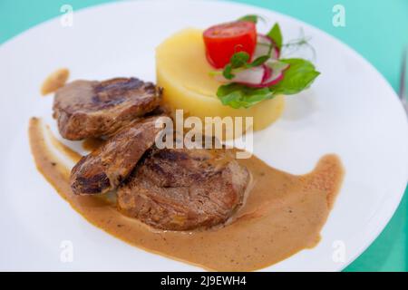 Fleischbällchen mit Kartoffelpüree auf einem weißen Teller. Fleisch in saurer Rahmsoße. Tomaten und Basilikum. Essen im Restaurant. Mittagspause. Medaillons. Stockfoto