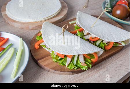 Zwei Tortillas gefüllt mit frischem Gemüse und Hühnerfilets auf einem Schneidebrett und einem Holztisch. Food-Trend. Modische Art der Verpackung Stockfoto