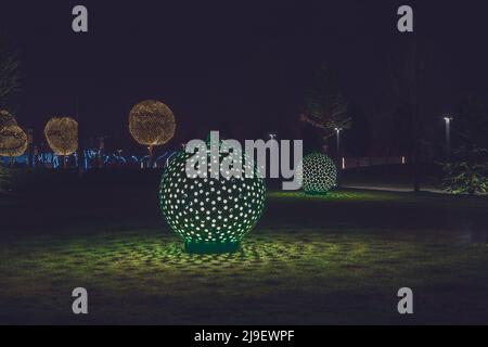 Russland, Krasnodar 07.01.2022. Runde glühende Kugeln werfen in der Nacht im dunklen Park gemusterte Schatten auf einen Rasen, der von glühenden Bäumen umgeben ist Stockfoto