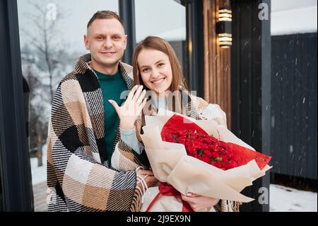 Freudige Frau, die einen Strauß Rosen hält und Hand mit Verlobungsring zeigt, während der Mann den Verlobten von hinten umarmt. Verlobt Paar in der Liebe stehen vor dem Haus am Wintertag. Stockfoto