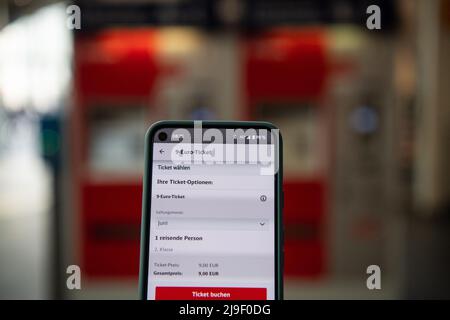 Bremen, Deutschland. 23.. Mai 2022. Die Buchungsseite für das 9-Euro-Ticket wird auf einem Handy mit einem Ticketautomaten im Hintergrund aufgerufen. Nach der Zustimmung von Bundestag und Bundesrat ist nun der Weg für das 9-Euro-Ticket frei. Ab Juni können die Verbraucher mit dem ÖPNV (ÖPNV) zu so günstigen Preisen in ganz Deutschland reisen. Das Ticket wird in den jeweiligen Apps, Fahrkartenautomaten und Kundenzentren aller Verkehrsunternehmen in Deutschland angeboten. Quelle: Melissa Erichsen/dpa/Alamy Live News Stockfoto