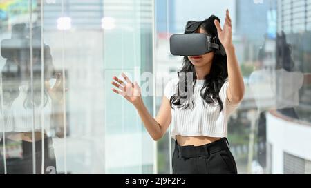 Schöne asiatische tausendjährige Frau, die ein VR-Headset trägt und im modernen Büro ein digitales Virtual-Reality-Spiel spielt. Stockfoto