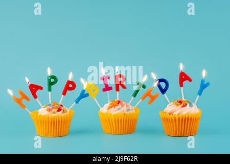 Drei Beerengeburtstag-Muffins in gelben Verpackungen mit bunten Kerzen auf blauem Hintergrund hochwertige Fotos für Kalender und Karten. Platz für Text Stockfoto