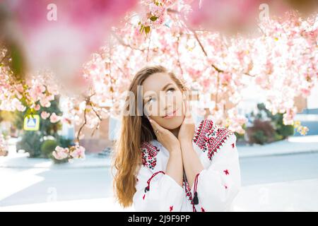 Stilvolle Frau in einem gestickten Hemd, die in der Nähe einer blühenden Sakura im Park posiert. Stockfoto