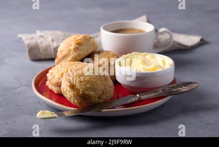 Hausgemachte, frisch gebackene Scones mit Käse und Kräutern auf einem roten Teller mit Butter, eine Tasse Kaffee auf einem grauen strukturierten Hintergrund. Traditionelles englisches Brötchen Stockfoto