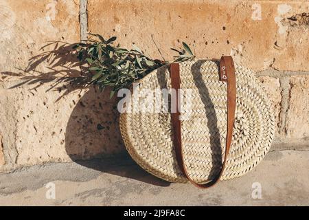 Sommerurlaub Stillleben. Runder französischer Strohkorb mit Ledergriffen und grünen Olivenbaumblättern und Zweigen im Sonnenlicht. Goldene Sandsteinmauer Stockfoto