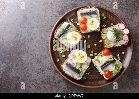 Offene Roggenbrot-Sandwiches mit Butter, Sprotte-Filet, Eiern, verschiedenen Gemüsesorten aus der Nähe auf einem Teller auf dem Tisch. Horizontale Draufsicht von oben Stockfoto