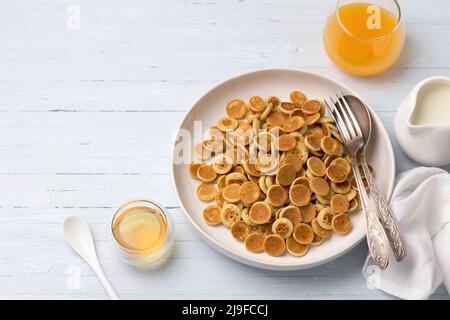 Teller Mini Pancakes Müsli mit Honig, Creme und frischem Orangensaft auf hellblauem Hintergrund. Trendige Küche. Hausgemachtes Frühstück. Draufsicht. Platz Stockfoto